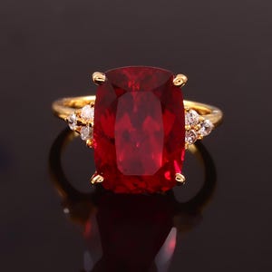 Può includere: Un anello d'oro con una grande pietra preziosa rettangolare rossa circondata da pietre preziose più piccole e trasparenti.