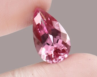 May include: A pear-shaped pink gemstone with a faceted cut. The gemstone is held in a hand.