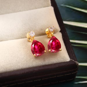 Puede incluir: Un par de pendientes de oro con piedras preciosas rojas en forma de lágrima y pequeñas piedras redondas transparentes.
