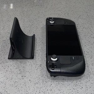 May include: A black handheld gaming device with a screen, buttons, and a joystick. The device is sitting on a grey countertop next to a black stand.