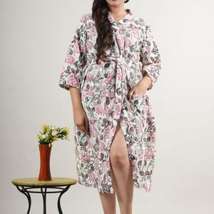 May include: A white floral print robe with pink and gray accents. The robe has a tie waist, pockets, and three-quarter sleeves. A small side table with a vase of flowers is in the background.