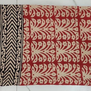 May include: Folded fabric with a red and beige floral pattern. A beige border with a black and beige geometric design runs along the left edge. The fabric is folded and appears to be made of a soft material.