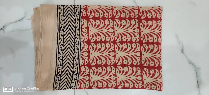 May include: Folded fabric with a red and beige floral pattern. A beige border with a black and beige geometric design runs along the left edge. The fabric is folded and appears to be made of a soft material.