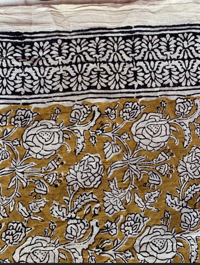 May include: A white cotton fabric with a mustard yellow floral print. The fabric features a black and white border with a repeating floral pattern.