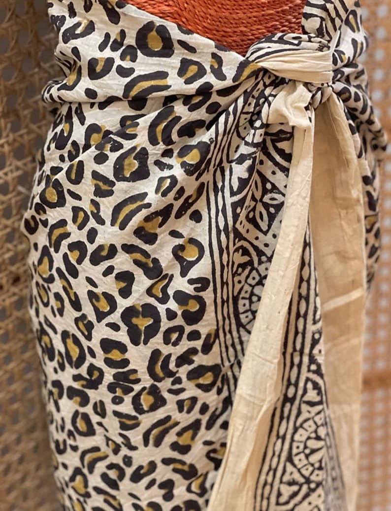 May include: A white cotton sarong with a black and gold leopard print design. The sarong is tied around the waist and has a white cotton trim.
