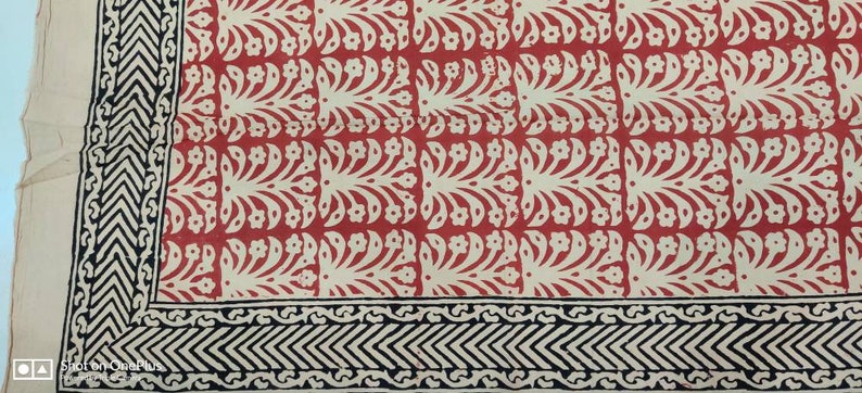 May include: A red and cream-colored textile with a repeating floral pattern. The border features a black and cream geometric design. The fabric appears to be a cotton or linen, suitable for home decor or apparel.