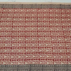 May include: A rectangular textile with a repeating pattern of red and cream-colored leaf-like shapes. The border is black with a geometric design. The background is a light beige color. This textile could be a tablecloth or a decorative fabric.