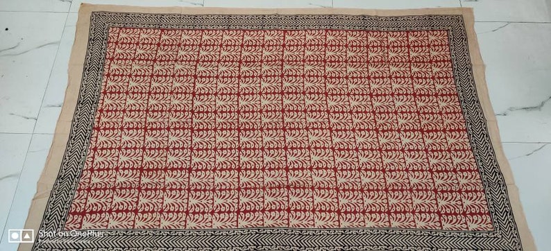 May include: A rectangular textile with a repeating pattern of red and cream-colored leaf-like shapes. The border is black with a geometric design. The background is a light beige color. This textile could be a tablecloth or a decorative fabric.