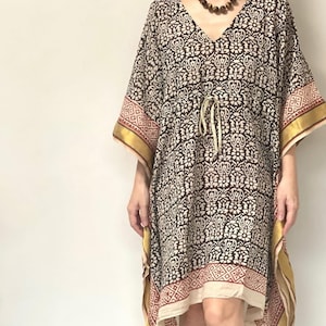 May include: A patterned, knee-length kaftan in black, cream, and red. The kaftan features a V-neck, wide sleeves, and a drawstring waist. Gold trim accents the sleeves and hem. A brown beaded necklace is also visible.