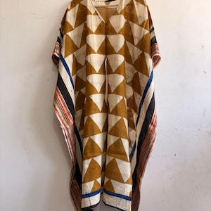 Könnte beinhalten: Ein gemustertes Kaftankleid in Beige, Braun und Weiß, das an einem Holzbügel hängt. Der Kaftan hat einen V-Ausschnitt und weite Ärmel. Die Kanten sind mit Streifen in Schwarz, Blau, Rot und Weiß verziert.