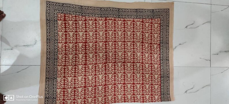 May include: A rectangular textile with a red and cream floral pattern. The design is arranged in a grid, with a black and cream border. The textile is likely a scarf or a decorative cloth.