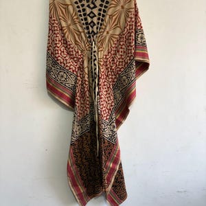 Puede incluir: Un caftán estampado en tonos beige, rojo y negro, colgado de una percha blanca. El caftán presenta un diseño geométrico y floral, con un cordón en el escote. Los bordes están ribeteados con un borde rosa y amarillo.