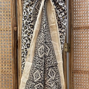 May include: A cream-colored sarong with a black and white geometric pattern. The sarong is tied around the waist and has a leopard print on the side.