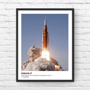 Peut inclure: Impression encadrée du lancement de la fusée Artemis II, avec une fusée orange vif et blanche sur fond de ciel bleu. La fusée est entourée d'un nuage de fumée blanche et de feu. Le texte en bas indique "Artemis II NASA Space Launch System (SLS) Rocket Launch 1er avril 2026."
