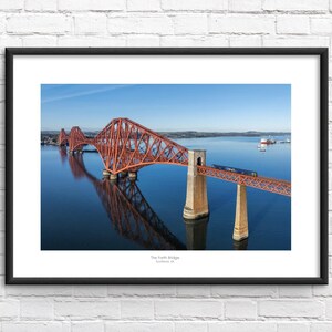 May include: Framed print of the Forth Bridge in Scotland. The iconic red bridge spans the water, with a train crossing it. The image is reflected in the calm, blue water below. The sky is clear and blue.