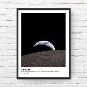 以下が含まれることがあります： 「Earthset」と題された額入りのプリントで、月の表面から昇る地球の写真が特徴です。この画像は、黒い空を背景に三日月型の地球を示しています。プリントには写真家の名前と日付が含まれています。
