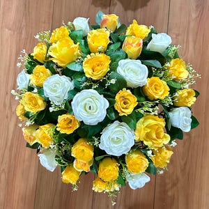 May include: A bouquet of artificial flowers featuring white and yellow roses. The roses are arranged in a circular shape and are surrounded by green leaves.