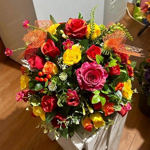May include: A colourful bouquet of artificial roses in shades of red, yellow, and pink, with green leaves and orange decorative netting. The arrangement is in a white wooden crate, creating a bright floral display.
