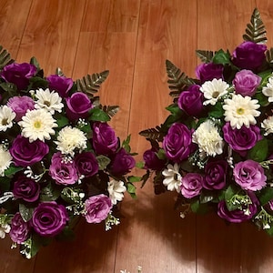 May include: Two floral arrangements with deep purple roses, white daisies, and green leaves. The arrangements are symmetrical, with a mix of textures and colours, creating a visually appealing display. The flowers are arranged in a compact, rounded shape.