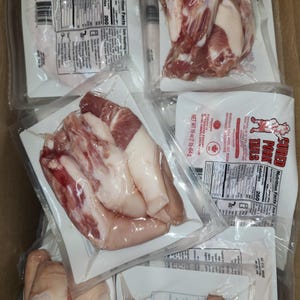 May include: Multiple packages of cured pork tails, each containing several pieces of meat, are stacked on top of each other. The packages are labeled with the text "Cured Pork Tails" and nutritional information. The packages are clear plastic and the meat is visible.