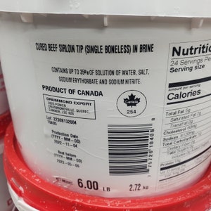 May include: A white plastic bucket with a red lid containing cured beef sirloin tip in brine. The label states "Cured Beef Sirloin Tip (Single Boneless) in Brine" and includes the product's weight, production date, and best before date. The label also states "Product of Canada" and includes a Canadian flag symbol. The nutritional information is listed on the right side of the bucket.