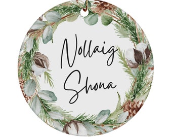 Könnte beinhalten: Runder Weihnachtsschmuck mit weißem Hintergrund und einem Kranz aus grünen Blättern, Tannenzapfen und Baumwolle. Der Text "Nollaig Shona" ist in schwarzer Schrift geschrieben. Ein goldener Faden ist zum Aufhängen befestigt.