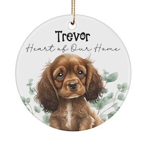 May include: A round ceramic ornament with a brown and white puppy with the text "Trevor Heart of Our Home" on a white background.