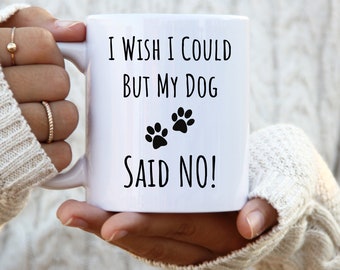 May include: White ceramic mug with the text "I wish I could but my dog said no!" in black, along with two black paw prints. The mug has a handle and is held by a person.