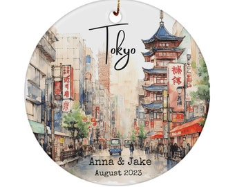 以下が含まれることがあります： 日本の東京の街並みを水彩画で描いた丸いセラミックオーナメント。オーナメントには、赤と青のアクセントが施された伝統的な日本の建物と、筆記体で「東京」の文字が描かれています。オーナメントの下部には「Anna & Jake 2023年8月」と記されています。