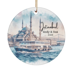 May include: A round ceramic ornament with a watercolor painting of the Hagia Sophia mosque in Istanbul, Turkey. The ornament features a ferry boat in the foreground and a bridge in the background. The text "Istanbul" is written in cursive at the top of the ornament, followed by "Andy & Sam 2023".