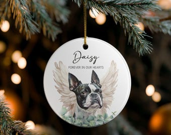 May include: A white ceramic Christmas bauble featuring a Boston Terrier with angel wings and eucalyptus leaves. The bauble has the name "Daisy" and the text "Forever in our hearts" in black script. A gold string is attached for hanging.