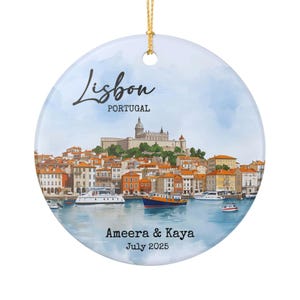 May include: Round ceramic ornament featuring a watercolor illustration of Lisbon, Portugal. The design includes the city skyline, boats, and the text "Lisbon Portugal". Personalized with "Ameera & Kaya July 2025". Includes a gold string for hanging.