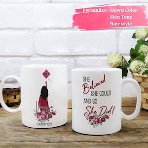 Puede incluir: Dos tazas de cerámica blanca con un tema de graduación. Una taza presenta a una mujer con una túnica granate lanzando un gorro de graduación al aire con el texto "Clase de 2023". La otra taza tiene el texto "She Believed She Could And So She Did!" con diseños florales.