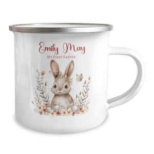 Pode incluir: Caneca de esmalte branco com borda prateada que apresenta uma ilustração em aquarela de um coelho marrom com uma coroa de flores rosa cercada por flores rosa e amarelas e folhas verdes. O texto "Emily May My First Easter" está impresso em rosa na caneca.