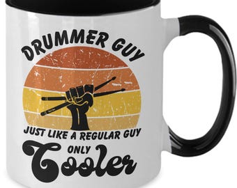 May include: White ceramic mug with a black interior, handle, and rim. The mug features the text "Drummer Guy" above a graphic of a hand holding drumsticks, set against a retro sunset design. The text "Just Like A Regular Guy Only Cooler" is below.