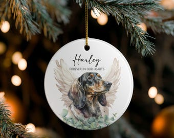 May include: A white ceramic Christmas ornament featuring a dachshund with angel wings and the name "Harley". The text "Forever in our hearts" is also displayed. The ornament hangs from a gold string.