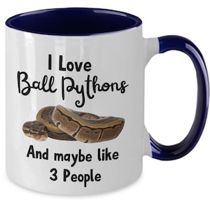 May include: White ceramic coffee mug with a blue rim and handle. The mug features a black and white graphic of a ball python and the text "I Love Ball Pythons And maybe like 3 People".