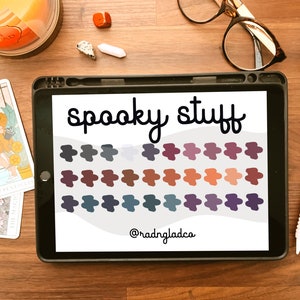 May include: An iPad displays the words "spooky stuff" above a grid of color swatches in shades of gray, purple, and orange. A tarot card, candle, and accessories are on a wooden surface.