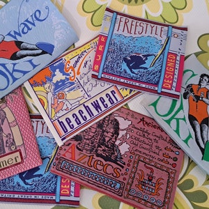 Puede incluir: Una colección de etiquetas rectangulares de tela para ropa con varios diseños gráficos, incluyendo un esquiador acuático, un surfista, un elefante y una ilustración de temática azteca, con texto impreso como Ski Wave, Freestyle y Summer.