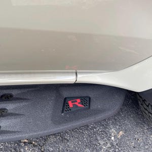 May include: A black running board with a red "R" emblem is attached to a light-colored vehicle. The emblem is rectangular and has a textured background. The vehicle's side panel and tire are also visible.