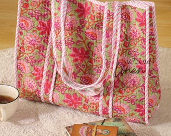 May include: A large tote bag with a floral pattern in shades of pink and green. The bag has white and pink striped trim and handles. A stack of books and a cup of tea are next to the bag.