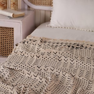 Puede incluir: Una manta color crema con un patrón geométrico negro de flechas y puntos. La manta está sobre una cama con una almohada blanca y una mesita de noche con un libro.