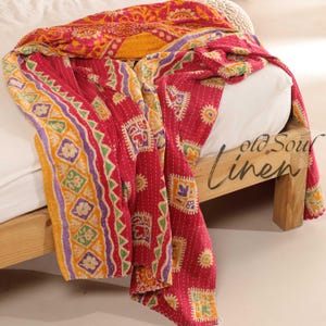 May include: A red, yellow and purple kantha quilt with a geometric pattern. The quilt is draped over a wooden bed frame.