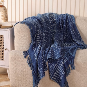 May include: A navy blue throw blanket with a white geometric pattern and fringe trim. The blanket is draped over a beige sofa, showcasing its intricate design and texture. The throw blanket adds a touch of elegance and comfort to any living space.