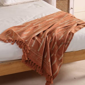Puede incluir: Una manta naranja con un patr&oacute;n geom&eacute;trico y flecos. La manta est&aacute; colocada sobre una cama, mostrando su textura y dise&ntilde;o. El patr&oacute;n consiste en l&iacute;neas blancas y naranjas. La manta est&aacute; hecha de un material suave.