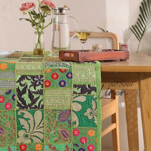 May include: A green patchwork table runner with floral and geometric patterns. The runner has a green border and is placed on a wooden table. A glass pitcher, a glass of liquid, and a wooden tray are on the table. A vase of pink flowers is also present.
