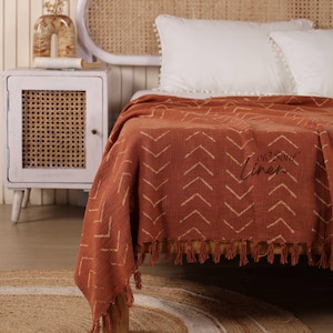 Op de afbeelding: Roestkleurige sprei met een crèmekleurig chevronpatroon en franjes. De sprei ligt op een bed met witte kussens. De tekst "old soul linen" is op de sprei gedrukt.