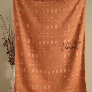 May include: A terracotta-coloured linen throw blanket with a repeating pattern of vertical lines and dots. The blanket has a fringed edge and the words "old Soul linen" are printed on the front.
