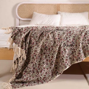 May include: A floral patterned throw blanket with a cream background and burgundy and green flowers. The blanket has a fringe trim and is draped over a bed with white pillows and a light-coloured wooden headboard.