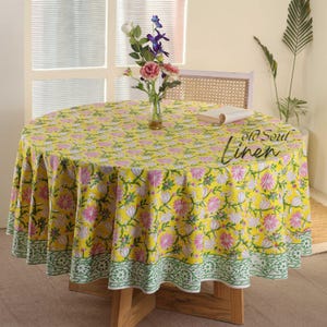 May include: A yellow round tablecloth with a pink and green floral print. The tablecloth has a green and white border. The tablecloth is on a round wooden table with a vase of flowers in the center.